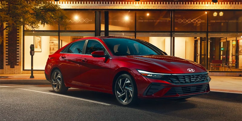 A shiny red Hyundai sedan parked on a street at dusk, storefront with illuminated windows in the background