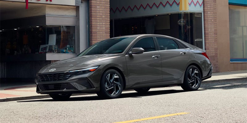 A dark gray Hyundai Elantra parked on a street in front of a brick building with storefront windows