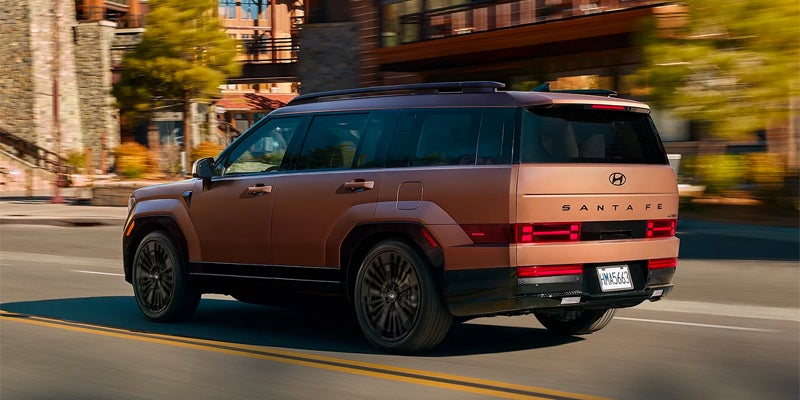 A bronze-colored Hyundai Santa Fe SUV drives toward the right on a paved road with buildings in the background