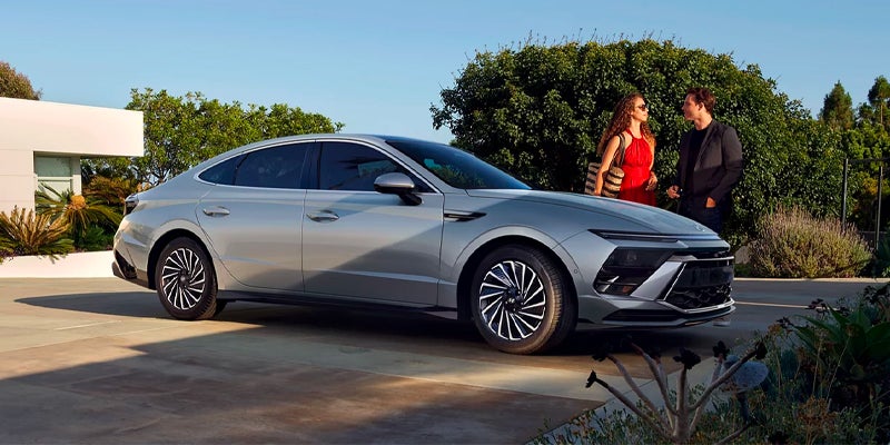 A silver modern sedan parked on a driveway with a couple standing nearby, in front of a white house