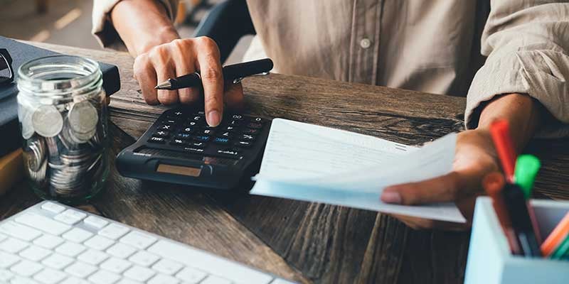 Hands calculating finances with a calculator and holding papers next to a jar of coins on a wooden desk