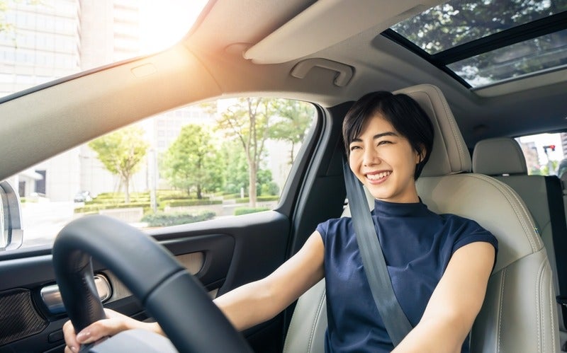 Smiling modern Asian woman with short hair, wearing a dark blue shirt and seatbelt, driving a car with a sunroof
