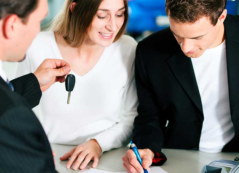 A sales person hands a smiling woman car keys as a man signs a document