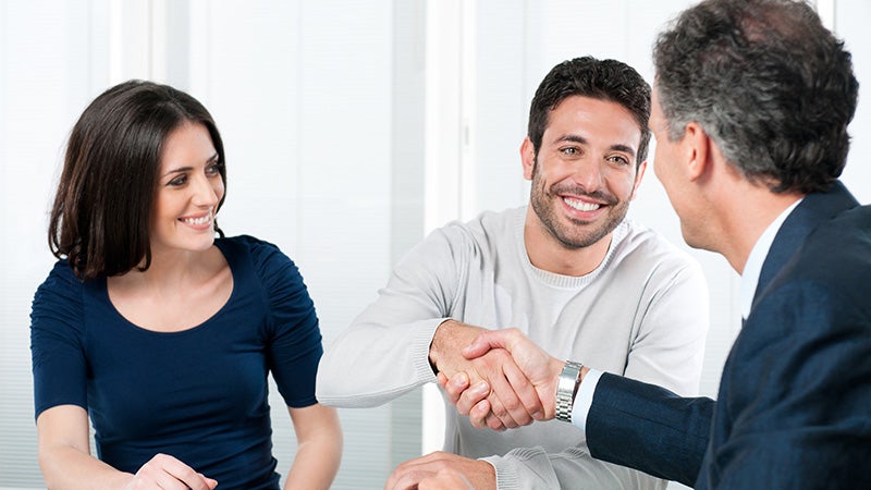 A car salesman hands keys to a happy woman as a man signs paperwork at a desk
