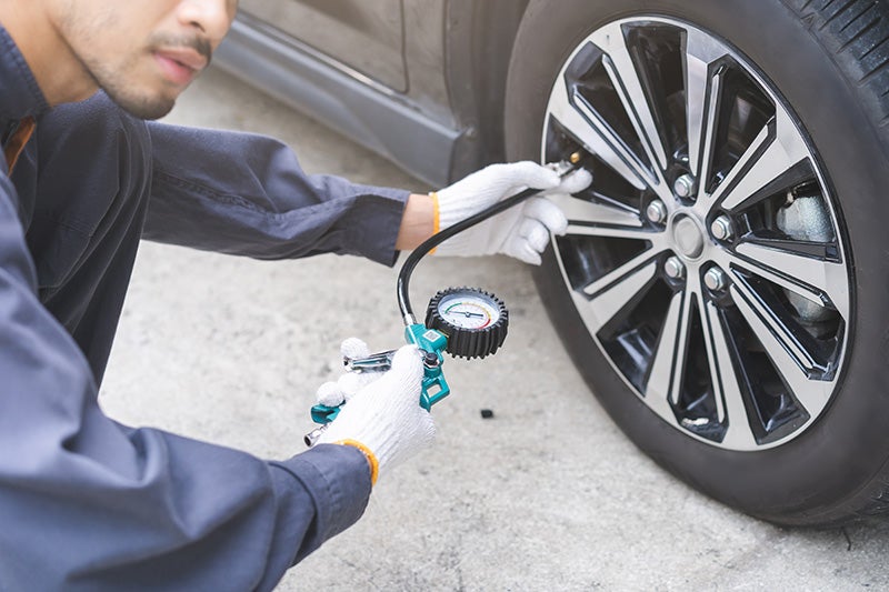 A man in work clothes and gloves uses a tire pressure gauge to check a car tire